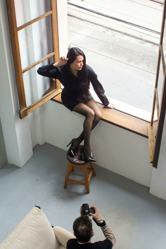 Indoor professional photo session with natural light through a large open window, featuring a model posing and a photographer in action.