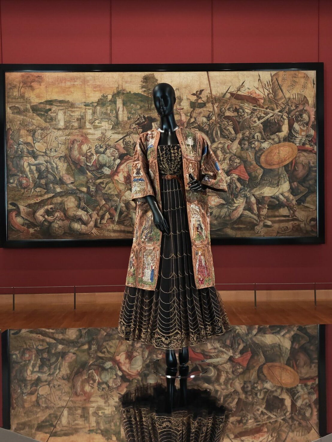 Mannequin de mode portant une robe plissée noire brodée d'or et un manteau richement orné, exposé devant un grand tableau de bataille classique dans un musée.